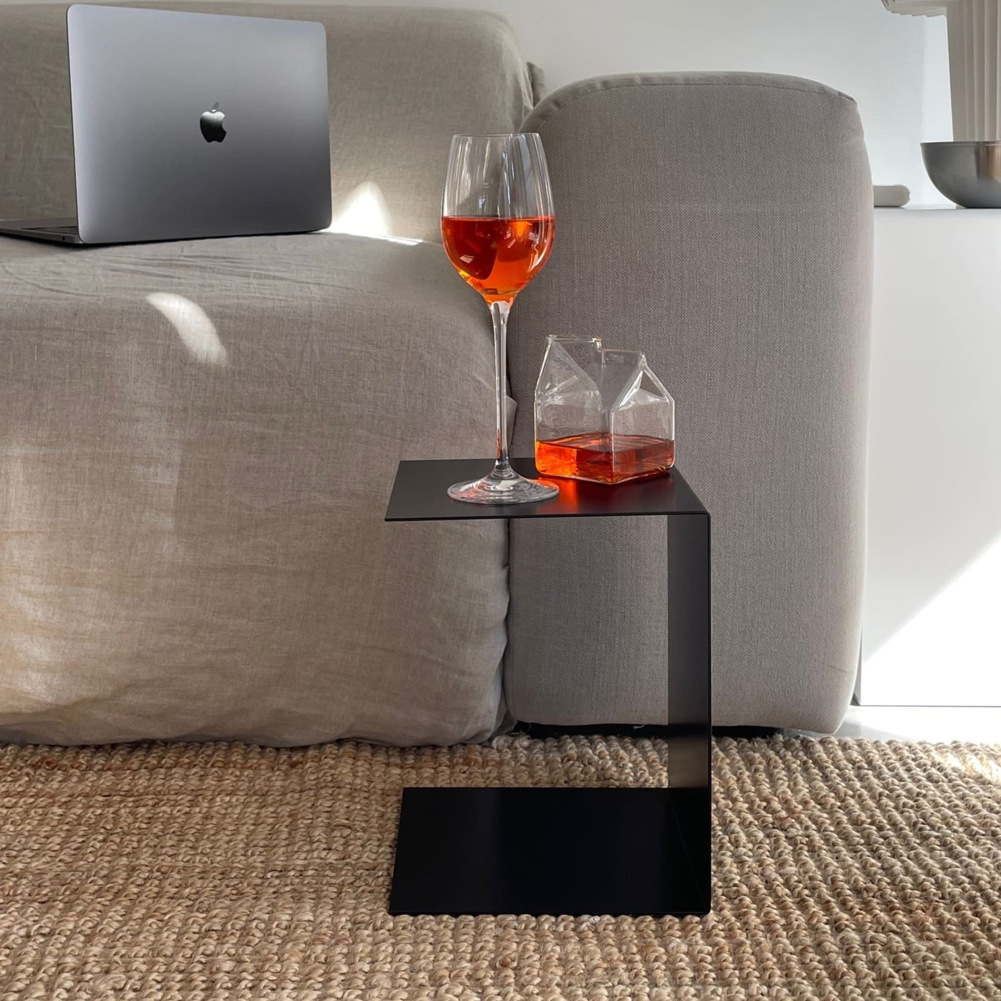 Kleiner schwarzer Beistelltisch mit Aperol-Glas und Karaffe, neben grauem Sofa auf geflochtenem Teppich