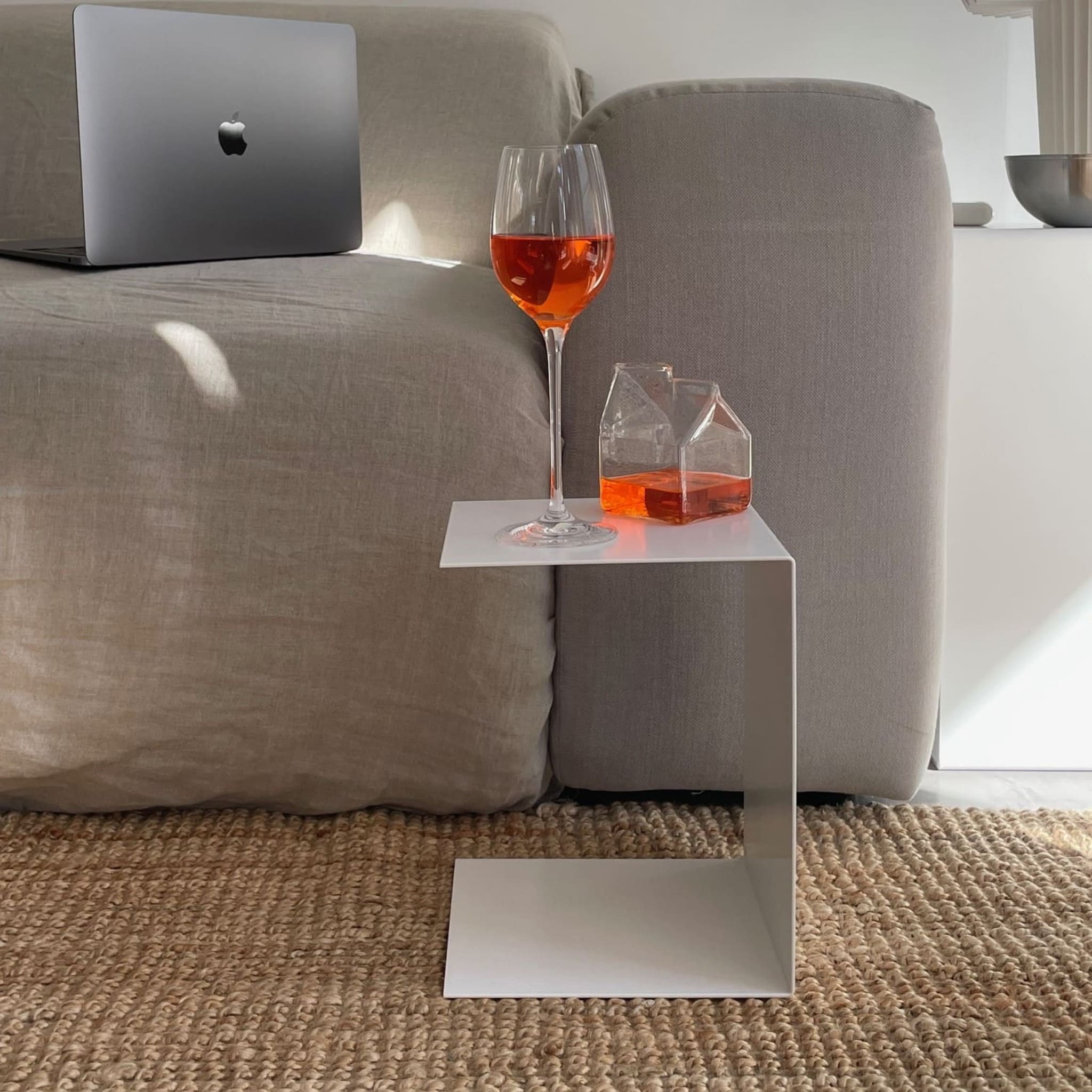 Kleiner weißer Beistelltisch mit Aperol-Glas und Karaffe, neben grauem Sofa auf geflochtenem Teppich